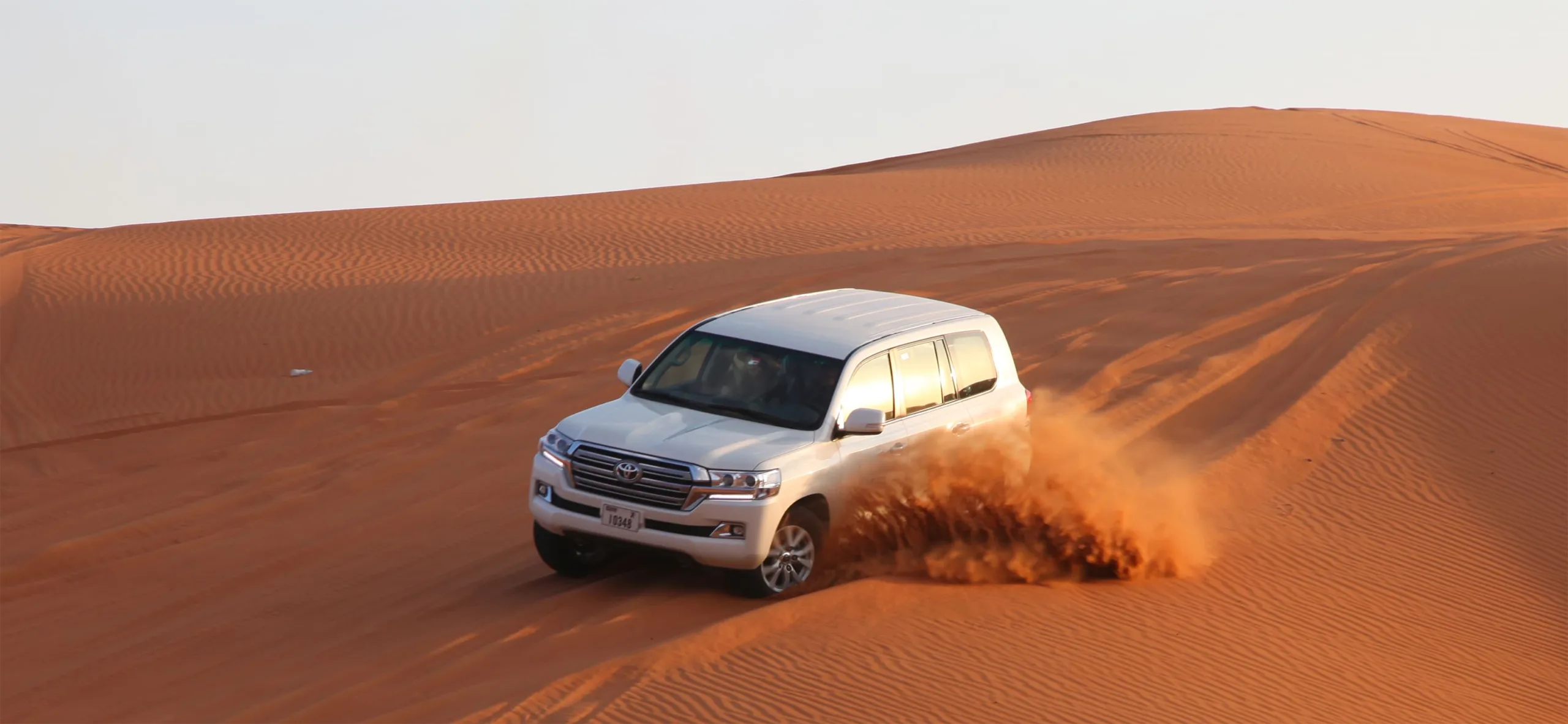 A white four-wheel-drive SUV performing a dune bash in the desert, with sand dramatically spraying from its tires