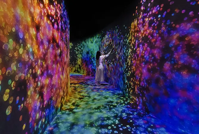 A person in a light-colored dress stands in a long, dark hallway with walls and floors that are projected with brilliant, colorful, and shifting light patterns, advertising teamLab Phenomena tickets.